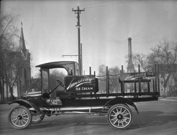 A brand new delivery truck parked outdoors near the Schoelkopf Automobile dealership (not shown) in the middle of East Washington Avenue. Two signs on the truck read: "American Ice Cream." In the background looking east are buildings lining both sides of the street, including a church building and a smokestack.