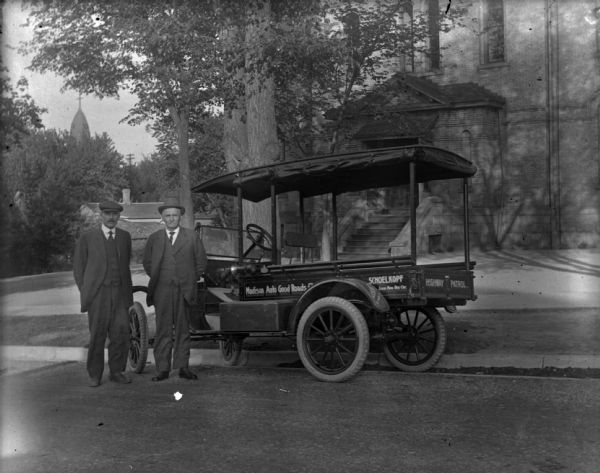 Two men in suits and hats pose outdoors near a Madison Auto Good Roads Highway Patrol vehicle parked at the curb. On the side of the truck are painted signs that read: "Madison Auto Good Roads ?," and "Schoelkopf Loans them this car." Building in the background is the Brayton School. Church in background is St. Patrick's.