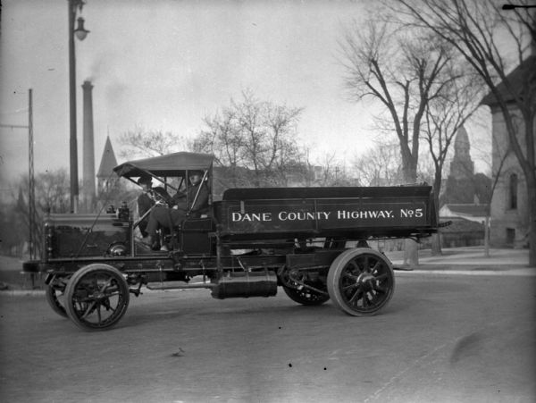 View looking east towards two men sitting in a Dane County Highway truck on East Washington Avenue. The truck is marked "No. 5" and is in front of the Schoelkopf Ford Automobile dealership (not shown) near the Madison Capitol Square. In the background is a smokestack and a church building.