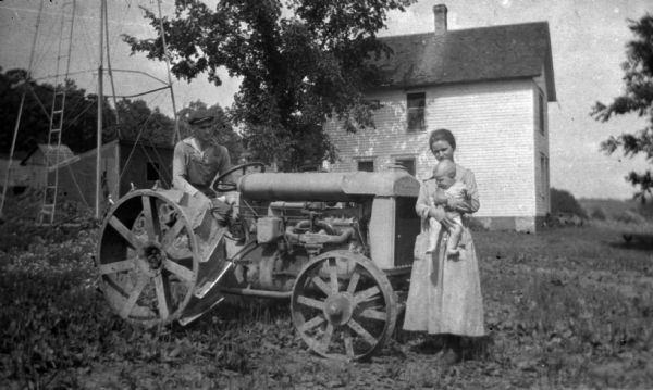 Group portrait of farm family. A man sits on a tractor on the left, and in front of the tractor on the right is a woman holding a baby. In the background is a metal support and ladder for what may be a windmill, and a clapboard house and other farm buildings.