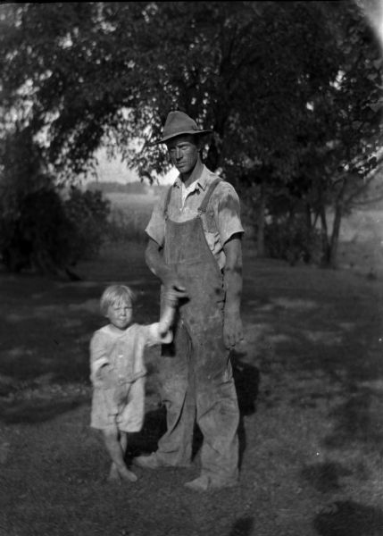 A farmer is holding a small boy by the hand while standing on a lawn. In the background are trees and shrubs.