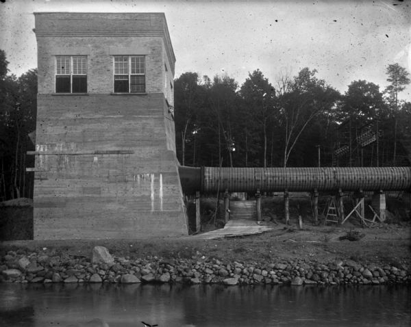 View across river or stream towards a pump house along a stony shoreline. A large pipe supported on trestles is attached to the right side of the building. Above the pipe are wires attached to a structure of what may be transformers. Ladders and lumber are under the pipe, as well as a wooden walkway.