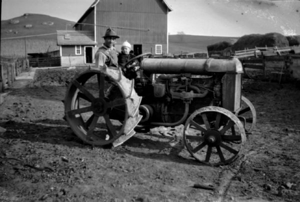 A man and child pose sitting on a tractor in a farmyard. In the background is a barn, and piles of hay behind a fence on the right. In the far background is a steep hill with rock outcroppings.
