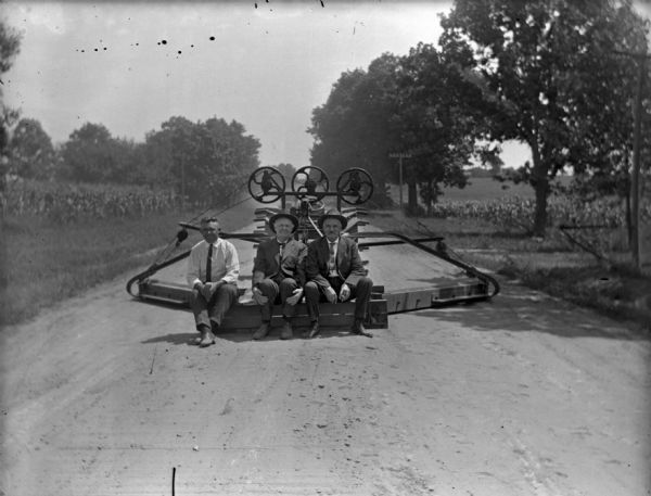 View down middle of unpaved road towards three men sitting on the back of a road grader in a rural area. Two of the men are wearing suits and hats, and the third man is in shirtsleeves.