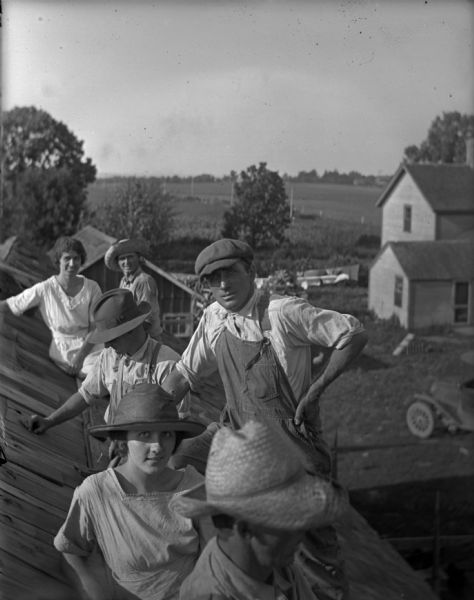 View along roof of four men in hats and work clothes, and two women in summer dresses posing on the side of the roof. One of the men is holding a hammer. In the background below is a farmhouse, a farm building and an automobile.