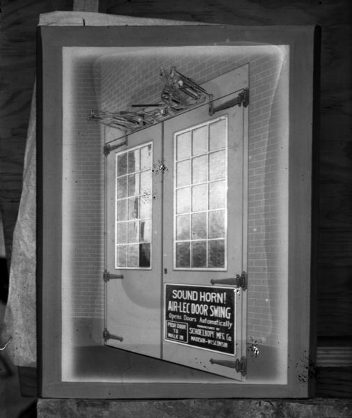 Framed display of a photograph or illustration of double doors with the Air-Lec automatic door opener system. The sign on the door reads: "Sound Horn! Air-Lec Door Swing Opens Doors Automatically, Manufactured by Schoelkopf Mfg. Co. Madison-Wisconsin," and "Push Door to Walk In."