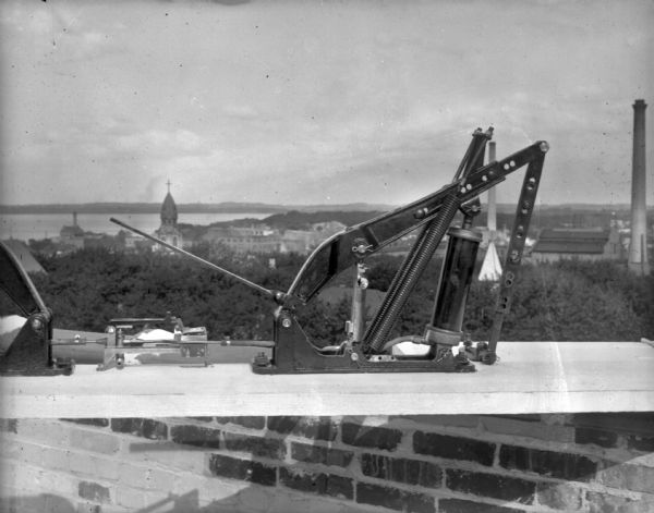 Outdoor view of an Air-Lec automatic door opener for swinging doors set up on a wood plank on a rooftop. Probably the top of Schoelkopf's manufacturing plant on N. Third Street. In the background below is a view of Madison with smokestacks, a church building, and Lake Mendota or Monona.