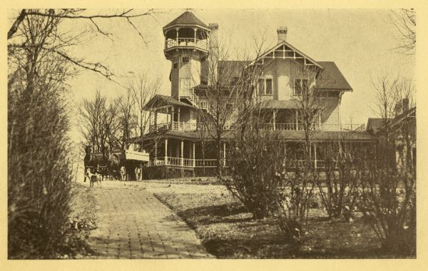View up brick path towards the exterior of the Conrad Seipp summer home at Black Point on Geneva Lake. A horse-drawn wagon is parked in front; a dog stands on the drive near the wagon. The family named the house "The Loreley" when it was built in 1888, but after 1921 simply used the name Black Point for the house as well as the property. The Queen Anne style house has a tall tower and multiple porches. The wing to the far right, referred to as the laundry house, served as a temporary dwelling for the Seipp family while the house was being built. It contained the kitchen and servants' quarters as well as laundry facilities.
