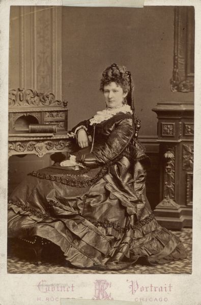 A full-length, seated cabinet card portrait of Catherine Orb Seipp wearing an elaborately detailed dress of heavy material. She is also wearing a bracelet, rings, drop earrings and a long chain necklace with a slide. A German-language book dated 1866 rests on the small desk beside her.