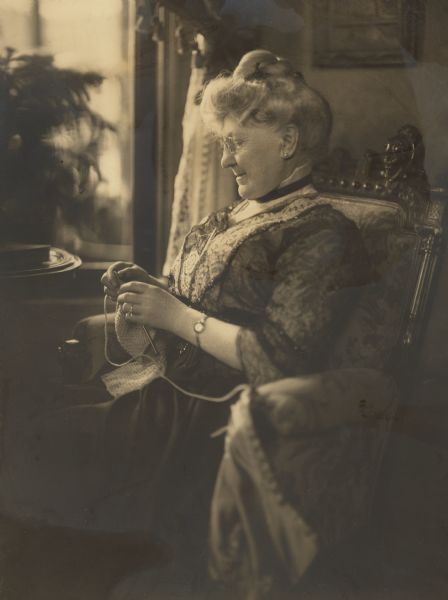 A seated portrait of Catherine Orb Seipp sitting near a window, knitting. Her upholstered chair has carved wood accents.