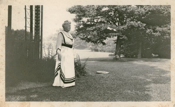 Catherine Orb Seipp stands on the lawn at Black Point, near one of the porches of the 1905 "cottage." She is wearing a summer dress with bold bands of dark trim. There is a trellis at the corner of the porch.