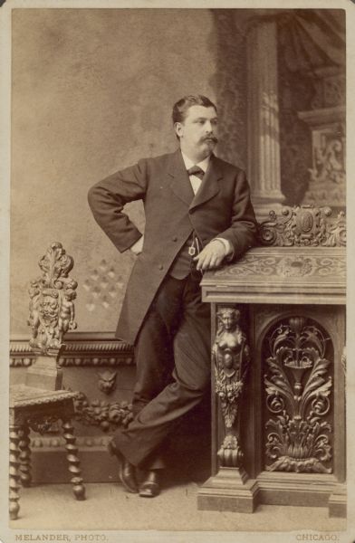 A standing full-length cabinet card portrait of William Conrad Seipp, the oldest son of Chicago brewer Conrad Seipp. He rests his left forearm on an ornate prop; there is an elaborately carved chair beside him.