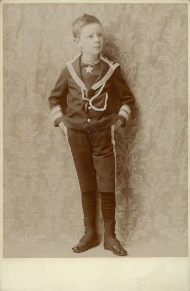 Conrad Seipp, Jr. (1883-1909), poses for a full-length portrait wearing a sailor suit in front of a tapestry background. He was the youngest child of Conrad and Catherine Seipp.