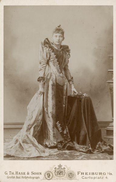 A full-length standing cabinet card portrait of Emma Sibylla Seipp (1868-1942), eldest child of Conrad Seipp with his second wife, Catherine Orb Seipp. She is wearing a highly tailored and detailed dress with a train. Her left hand rests on a book on a small table. Members of the Seipp family traveled regularly to Germany, where this photograph was taken.