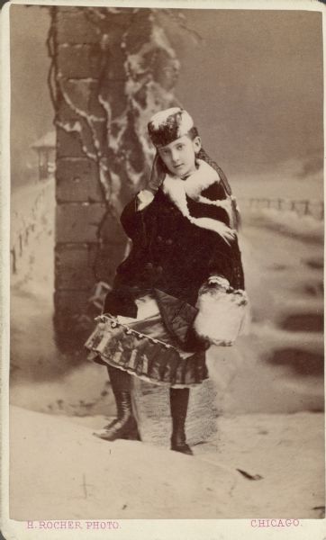 A standing, full-length studio portrait of Clara Seipp, a daughter of Chicago brewer Conrad Seipp. She is dressed in a dress and coat with hat, muff and gloves; she wears high boots and her hair is in a long braid.  Painted backdrop depicts snow-covered landscape.