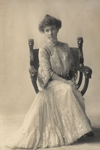 Full-length portrait of Alma Seipp posing seated in an ornately carved chair. She wears a floor-length gown with high collar and ribbon trim. Alma is a daughter of Conrad Seipp, the prominent Chicago brewer and builder of Black Point Estate.