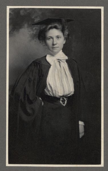 A standing, three-quarter length portrait of Alma Seipp wearing a graduation cap and gown posed in front of a painted backdrop. She has her hand on her hip and the gown is open to reveal her skirt and shirtwaist. Alma graduated from Wellesley College in 1899.