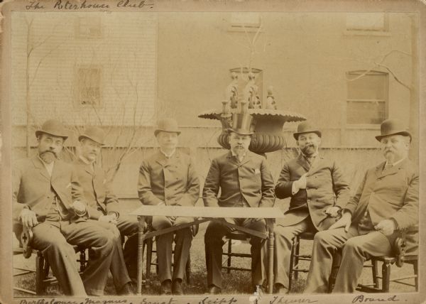 Six prominent German American brewers pose outdoors, seated around a table, for a portrait labeled "The Poterhouse [sic] Club." Well-dressed in suits and hats, the men are identified by last name. They include, from left, Philip Bartholomae, president of Bartholomae and Leicht Brewing Co. of Chicago; Christian Magnus, owner of Eagle Brewery and Bottling Works of Cedar Rapids, Iowa; one of the three Ernst brothers (C. Emil, Leo or Otto) of the Ernst Brothers Brewery of Chicago; Conrad Seipp, founder and president of Conrad Seipp Brewing Co. of Chicago; Joseph Theurer, owner of Schoenhofen Brewing Co. of Chicago, and Michael Brand of Michael Brand Brewing Co., Chicago. Seipp wears a top hat; the others wear fedoras. There is a large cast iron urn behind Seipp.