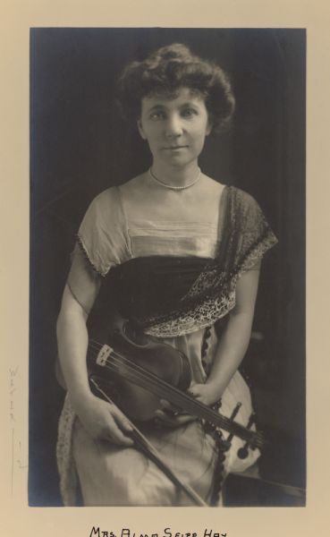 A seated, three-quarter length portrait of Alma Seipp Hay, a daughter of Chicago brewer Conrad Seipp. She is dressed in a finely detailed gown and holds a violin and bow. She is wearing a single strand of pearls.