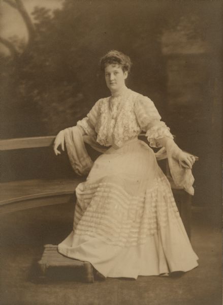 A seated, full-length portrait in front of a painted backdrop of Elsa Seipp, daughter of Chicago brewer Conrad Seipp. She is wearing a floor length gown with full skirt and ribbon trim. The dress is tailored with a very narrow waist. Her accessories include a light shawl and a pendant on a long chain.
