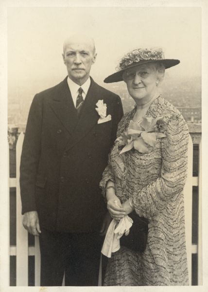 Albert and Elsa Seipp Madlener pose for their portrait on the day of their son Otto's wedding to Virginia Wetmore at the Cathedral of St. John the Divine. They are standing in front of a picket fence at an elevated location on the upper west side of New York City. Albert wears a suit and boutonnière; two orchids make up Elsa's corsage.