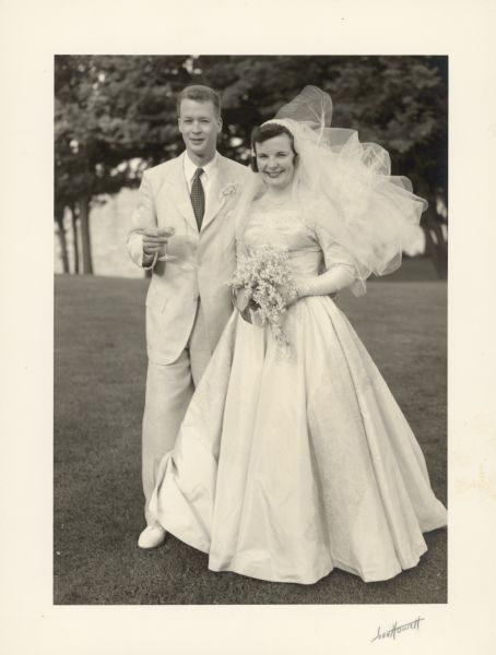 Claire Chapin, granddaughter of Clara Seipp Bartholomay, with her husband, William Frederick Carr, on their wedding day at Black Point Estate. Geneva Lake is visible in the background. The bride is wearing a wedding dress with full, floor length skirt and waist length veil; the groom is in a summer suit and light-colored shoes. He is holding a champagne glass. The bride is holding her bouquet.