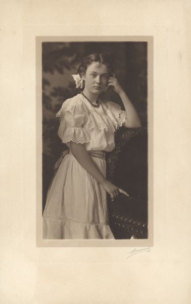 A standing, three-quarter length studio portrait of Alma Schmidt at age 12. She is wearing a summer dress with cut-work decoration. She has a bow in her hair and is leaning against the elaborately carved back of a chair.