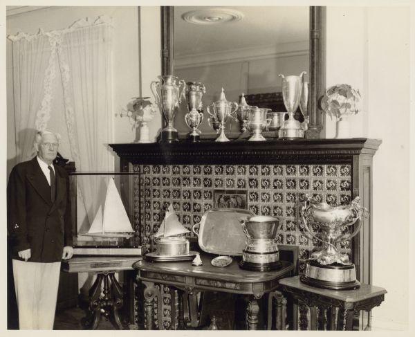 Ernst Conrad Schmidt, wearing conventional yachting attire of dark blazer and light-colored trousers, poses with trophies won by his boat, the "Senta III." The trophies are displayed on tables and the mantle in the living room at Black Point. There are decorative tiles on the fireplace surround. The trophies on the mantle were awarded by the Lake Geneva Yacht Club; most of the others were awarded by the Inland Lake Yachting Association.