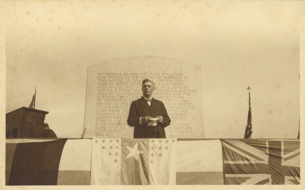 Dr. Otto L. Schmidt delivers a speech from a platform in front of a stone monument on which is inscribed the text of Abraham Lincoln's Farewell Address to Springfield, Illinois. Schmidt, in his capacity as the president of the Illinois State Historical Society and chairman of the Illinois Centennial Commision, was participating in the dedication of Andrew O'Connor's bronze sculpture of Lincoln which stands on the opposite side of the monument. The official Illinois Centennial Banner, center, and other flags are draped in front of the platform; a United States flag stands at the right.  There is an upright piano on the left.