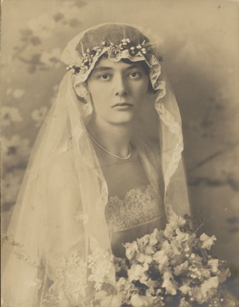 Alma Schmidt poses for a waist-up portrait on the day of her wedding to William F. Petersen. She holds her bouquet of sweet peas and lilies of the valley. Her dress and veil have lace trim.