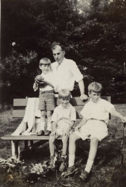Dr. William Petersen stands behind a bench posing with his three sons, posed on bench, from left, William Otto (standing, holding a ball), Conrad William, and Edward Schmidt Petersen. The boys are wearing shorts and light-colored shirts. Written on the reverse of the photograph is "Sunday - August 10, 1930 You can see how the children needed hair cuts. We are always so busy that there is never time for that. However last [illegible]."
