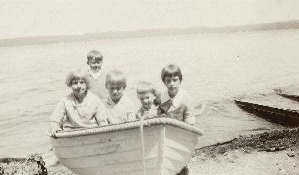 The three sons (center) of Dr. William and Alma Schmidt Petersen pose in a row boat on the shore of Geneva Lake with two unidentified girls. The boys are, from rear, Conrad William, Edward Schmidt, and William Otto Petersen. There are boat tracks on the right, and the far shoreline is in the background.