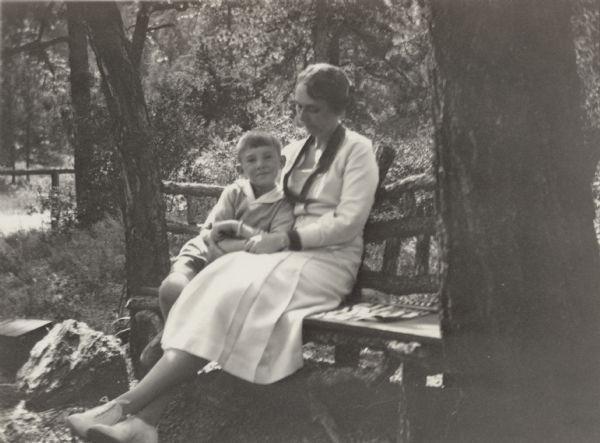 A smiling William Otto Petersen poses with his mother, Alma Schmidt Petersen on a rustic bench at Green Mountain Falls, Colorado. William is wearing shorts, a sweater and shirt. Alma is wearing a light-colored dress with dark collar and cuffs.