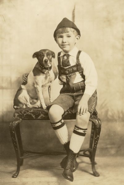 William O. Petersen, in a costume (Tracht) of Bavarian or Tyrolean style, poses seated on a bench with his arm around a small dog. He is wearing short lederhosen, long stockings, and a cap with a feather. His necktie and suspenders are decorated with edelweiss blossoms.