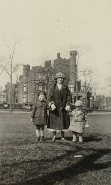 Edward, left, and Conrad Petersen, right, pose while holding hands with an unidentified young woman standing between them. They are on a lawn in front of the Potter Palmer mansion at 1350-60 North Lake Shore Drive. The boys lived with their parents, William F. and Alma Schmidt Petersen, a block away at 1322 North Astor Street. There are cars on a street in the background. The Palmer mansion, built in 1882, was faced with granite quarried near Endeavor, Wisconsin. It had a massive chimney and multiple crenellated towers. The mansion was demolished in 1950.