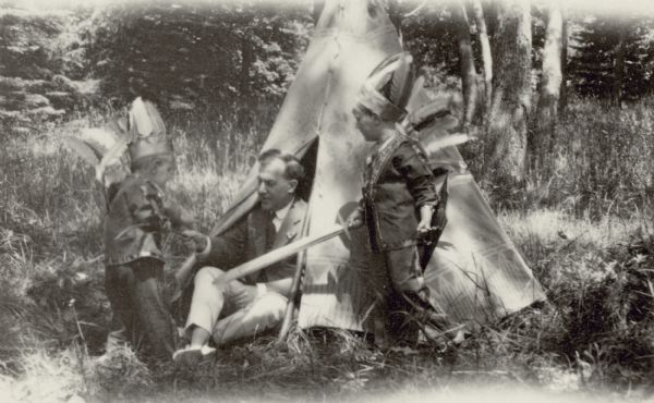 Dr. William F. Petersen sits in the doorway of a toy teepee playing with his sons Conrad, left, and Edward, dressed in Indian costumes. Edward wields a toy sword. Another child is obscured by Conrad.
