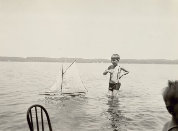 Edward Schmidt Petersen sails a toy boat in the shallow waters near the shore of Geneva Lake at Black Point. There is a chair and another child in the foreground.