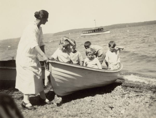Alma Schmidt Petersen, standing on the shore of Geneva Lake at Black Point, steadies a rowboat with her three sons and two other boys in it. Conrad Petersen stands in the bow, William Otto faces the camera, center, and Edward is at far right. There is a passenger launch out in the lake, and the far shoreline is in the background.