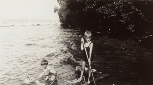 Conrad William Petersen poles a raft in the shallow water of Geneva Lake along the shore at Black Point. His younger brother, William Otto Petersen rides along. The Black Point pier is in the background.