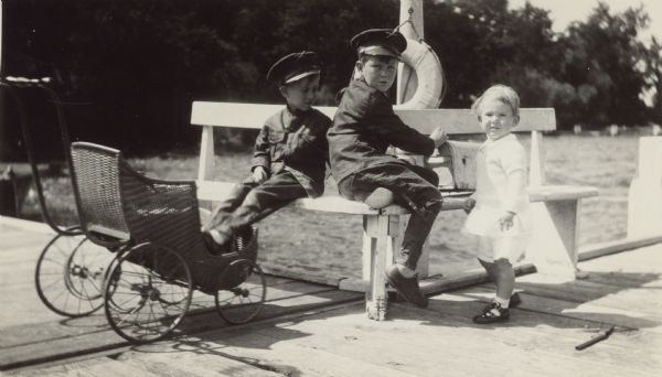 Conrad, resting his feet in a baby carriage, and Edward, center, sit on a bench on the pier at Black Point; their younger brother William stands at right. Edward has his hand on the top of a mailbox sitting on the bench beside him. A (toy?) gun is lying on the pier near William. Conrad and Edward wear water resistant jackets and caps. There is a life preserver on the post behind the bench. On the reverse of the photograph is written "Regatta Aug. 1928."