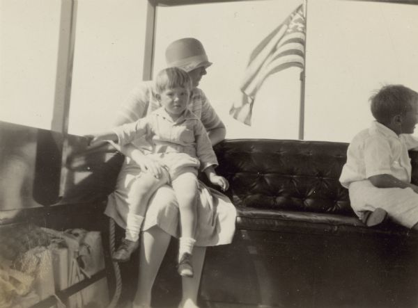 William Otto Petersen sits on his mother's lap as they ride inside a passenger launch on Geneva Lake. One of William's older brothers sits on the right. The seat is nicely upholstered and there is a flag outside at the stern.