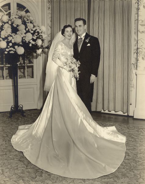 Conrad William and Kathryn Anne (Reebie) Petersen pose for a full length portrait on their wedding day in Winnetka, Illinois. The bride wears a long satin gown with train and three-quarter length veil; she holds a bouquet of stephanotis blossoms. The groom is in a traditional morning suit with striped trousers. He has a pearl stick pin and lily of the valley boutonniere.  There is a large floral display of chrysanthemums in a floor vase on the left.