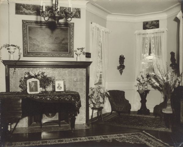 A grand piano with ornate shawl stands in front of the fireplace in the music room at Black Point. The fireplace surround is covered with ornate ceramic tiles. There are oriental rugs on the wood floors and lace curtains hang in the windows in a bay. There are painted friezes below the crown molding over the fireplace and one of the windows. The room is decorated with potted plants and cut flowers. Framed photographs of Catherine Seipp (left) and one of her daughters stand on the piano. There is a landscape painting over the fireplace and a simple six arm chandelier hangs from the ceiling. On the reverse of the photograph is written, "At the time of Aunt Clara and Uncle Henry's 60th wedding anniversary." Clara Seipp, daughter of Chicago brewer Conrad Seipp, married Henry Bartholomay, Jr. July 12, 1894.