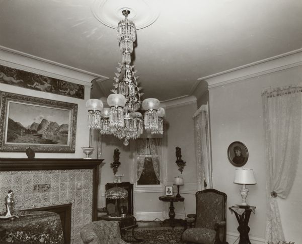 An ornate chandelier with gas light style shades and many prisms dominates a photograph of the music room at Black Point. There is a painted frieze below the crown molding over the fireplace just above a large landscape painting. The fireplace surround is covered with decorative tiles. There are lace curtains at the windows in the bay at the end of the room. A lamp with a leaded glass shade is on a small table.