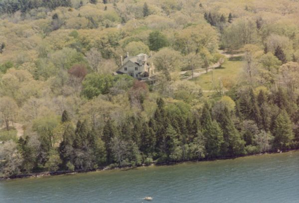 A small boat is seen on the waters of Geneva Lake in this aerial view of Black Point Estate. The main house with its distinctive tower and porches is easily seen above the trees.