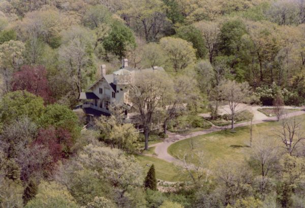 This aerial view of the main house at Black Point Estate shows the brick drive and walkways near the house. Trees are just budding out.  The house's distinctive tower and porches are also seen. The shore of Geneva Lake is out of view, to the left of the house.