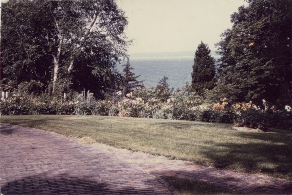 A view from near the main house at Black Point Estate shows the brick drive and walk in the foreground.  Geneva Lake is seen in the background, framed by a flower garden in full bloom and large trees.