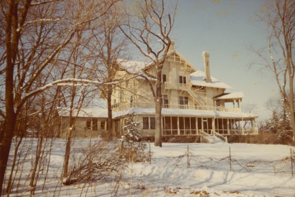 A winter view of the northeast side of the main house at Black Point Estate. There are two large chimneys, and first and second floor porches. A portion of the first floor porch is screened. Vines are growing on a low fence along a drive in the foreground. There is a small single story addition at the rear where a two-story attached "laundry house" originally stood. The house, overlooking Geneva Lake, was built by Chicago brewer Conrad Seipp in 1888 and initially named the Loreley.
