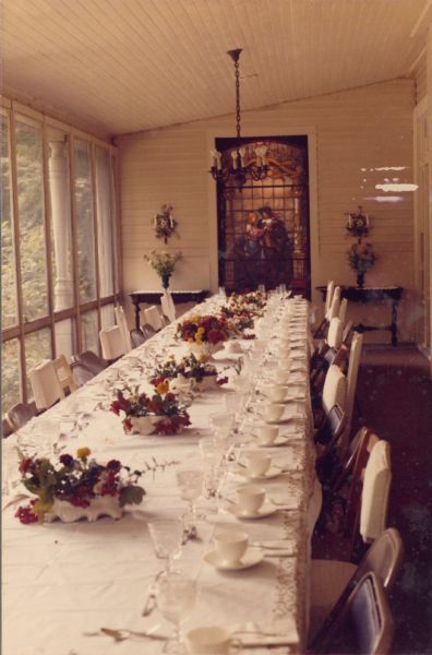 Low flower arrangements decorate a table set for at least 25 people on the screened dining porch at the main house at Black Point Estate.  There is a large stained glass window set into the far wall. The window was moved to Black Point from one of the Seipp family mansions in Chicago. The house, overlooking Geneva Lake, was built by Chicago brewer Conrad Seipp in 1888 and initially named the Loreley.