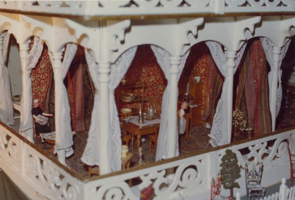 A close-up view of the kitchen of an antique doll house at Black Point Estate shows partial exterior walls of fretwork and columns decorated with draperies. There is miniature tableware and furniture including a table, shelves and cupboard. A doll in a maid's uniform is on the left and a child doll is seated at the table.